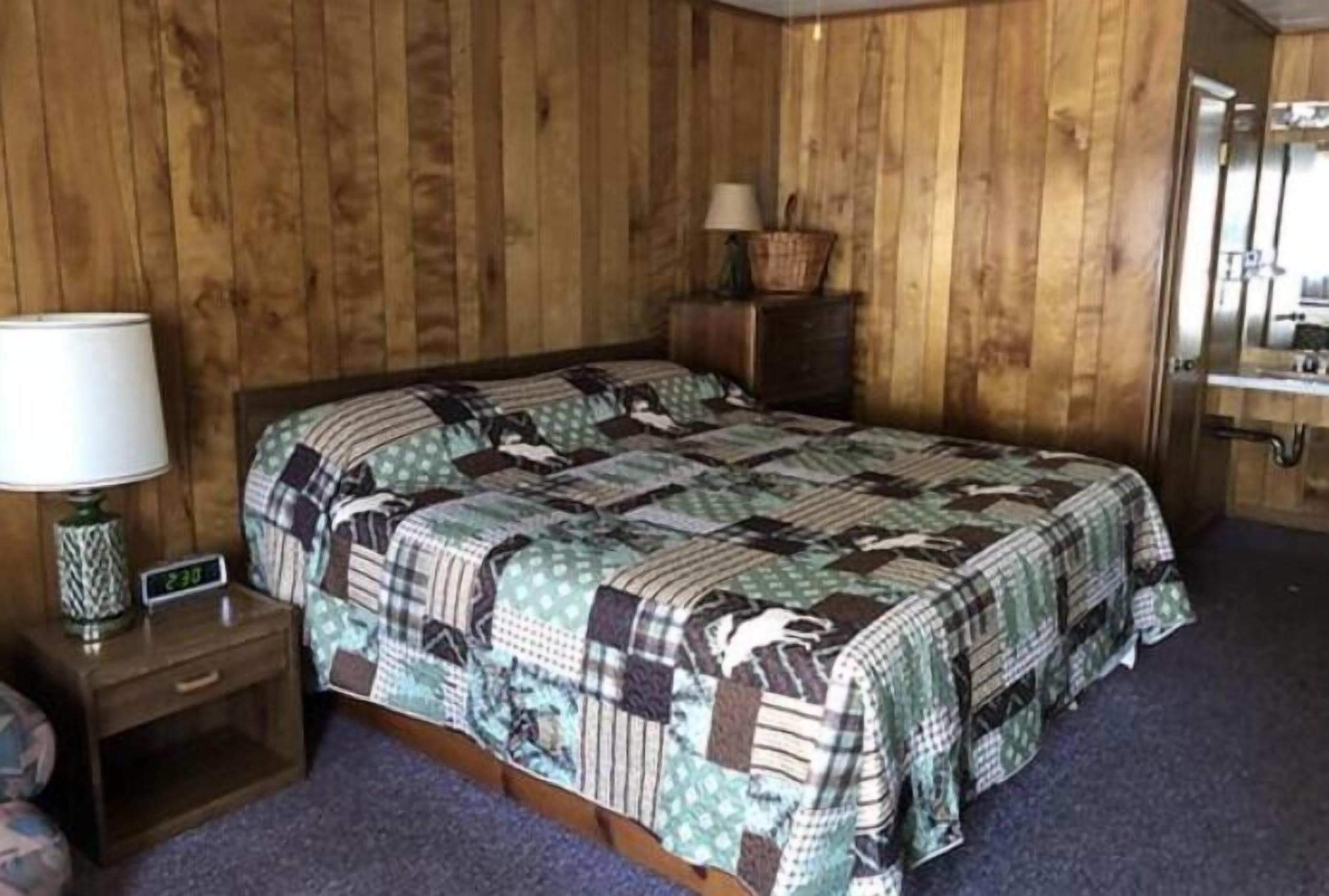 Una habitación con paneles de madera, una cama, una mesita de noche con una lámpara, un aparador y un espejo.