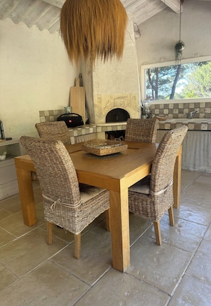 Une table à manger en bois, des chaises en osier, un luminaire suspendu et un coin cuisine avec un mur recouvert de carreaux et une fenêtre.