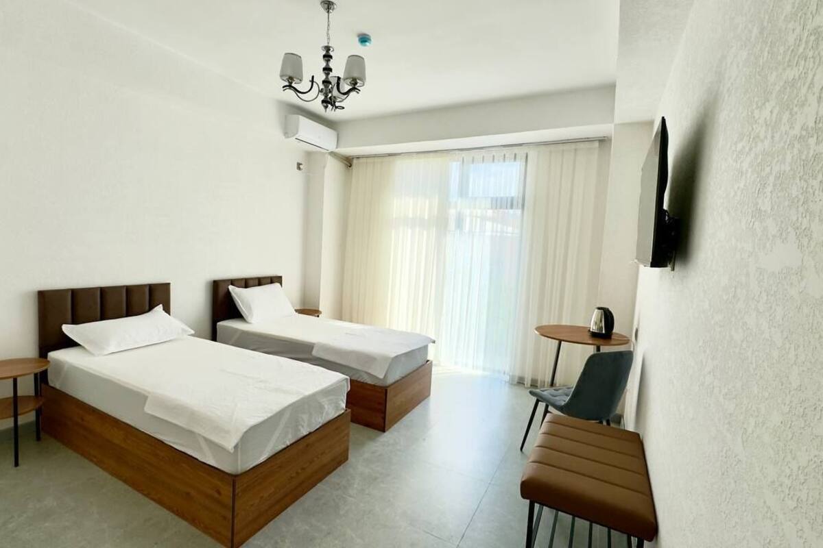 Habitación de hotel con dos camas, una mesita, una silla, televisor montado en la pared y ventana con cortinas.