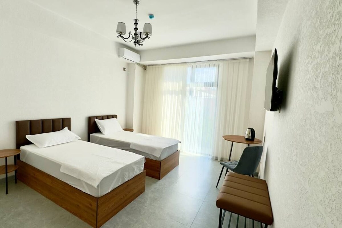 Quarto de hotel com duas camas, uma mesinha, uma cadeira, televisão presa na parede e janela com cortinas.