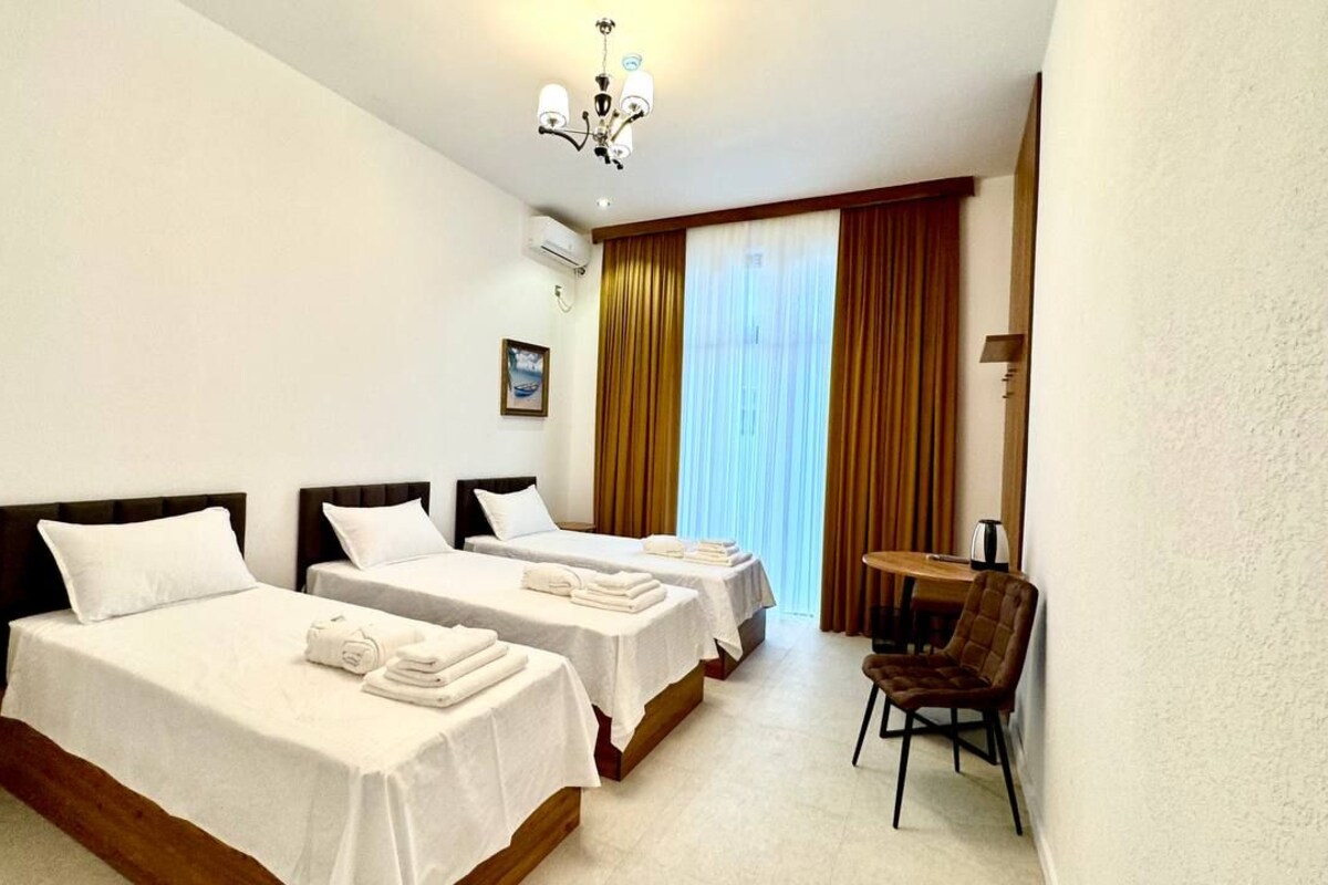 Una habitación de hotel con dos camas, una mesita, una silla y un ventanal con cortinas.