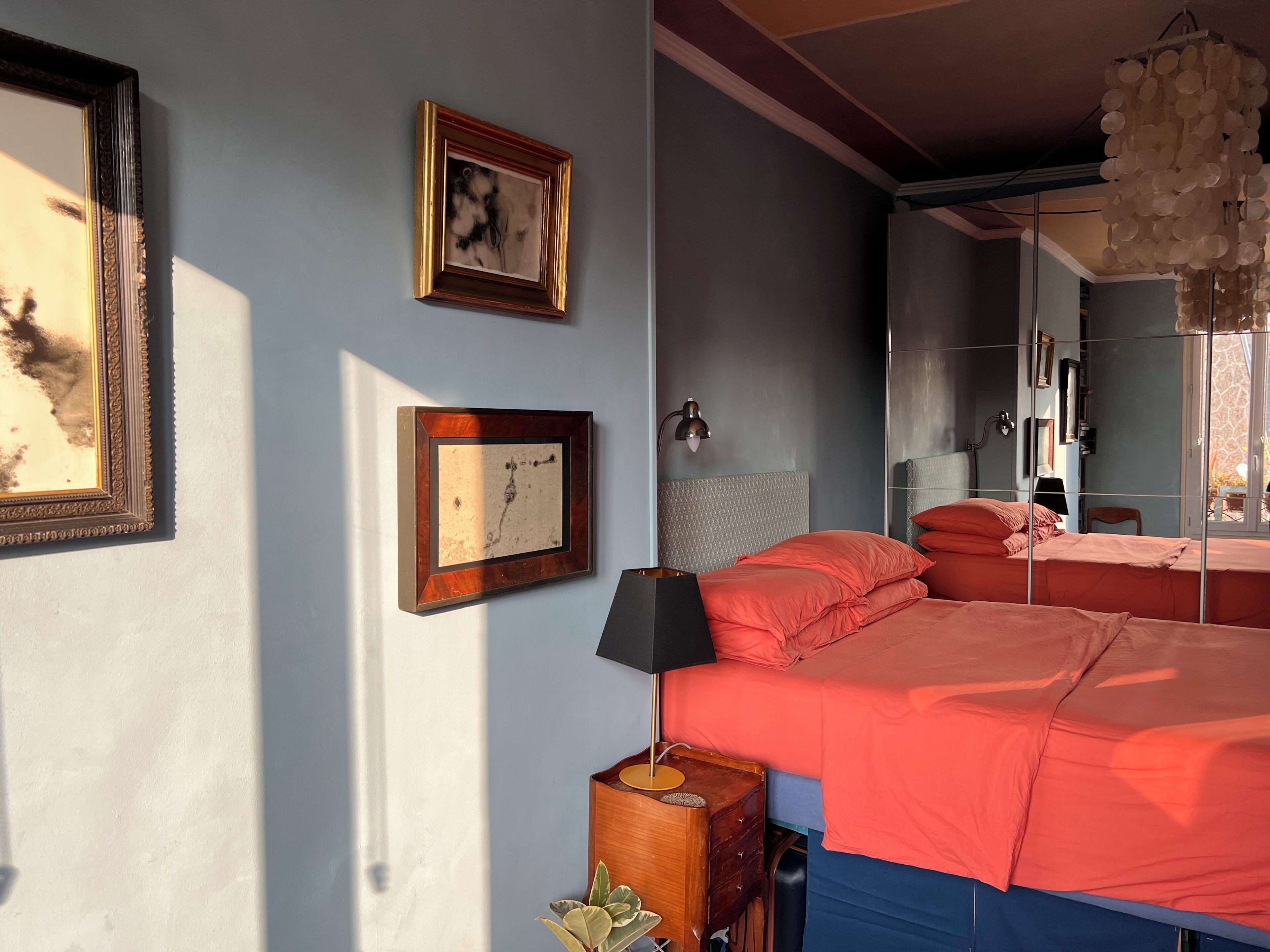 A bedroom with a bed, two framed pictures, a nightstand, a lamp, and a hanging light fixture.