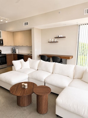 A modern living room with a sectional sofa, two round wooden coffee tables, a kitchen area with wooden cabinets, and a dining table with chairs.