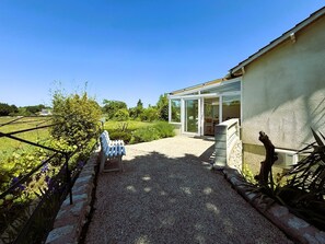 Una casa amb un carril de grava que condueix a una entrada amb porta de vidre, envoltada d'un jardí amb diverses plantes i una bosc.
