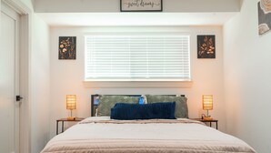 Een slaapkamer met een bed, twee bedlampen, een raam met jaloezieën en twee aan de muur bevestigde foto's.