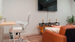 Een moderne woonkamer met een flatscreen televisie, een witte bureaustoel, een bruine bank, een klein tafeltje met een plant en een decoratieve plank met trofeeën.
