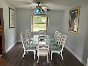 A dining room with a glass table, white chairs, a ceiling fan, and a window with a view of greenery.