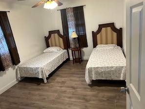 A room with two single beds, a small table, a lamp, and a ceiling fan.