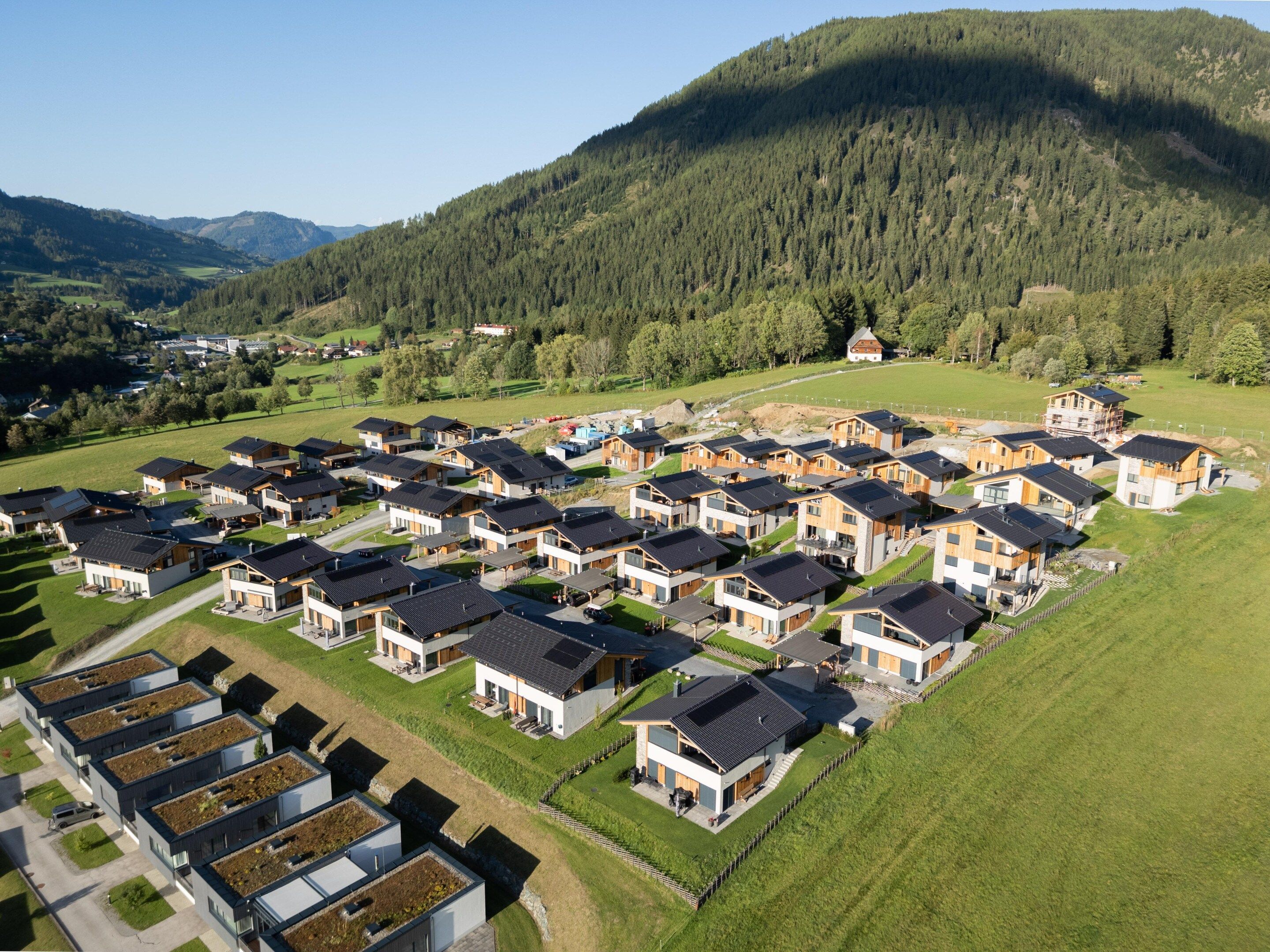 Un barrio residencial con casas modernas dispuestas en filas, rodeado de campos verdes y una colina arbolada.