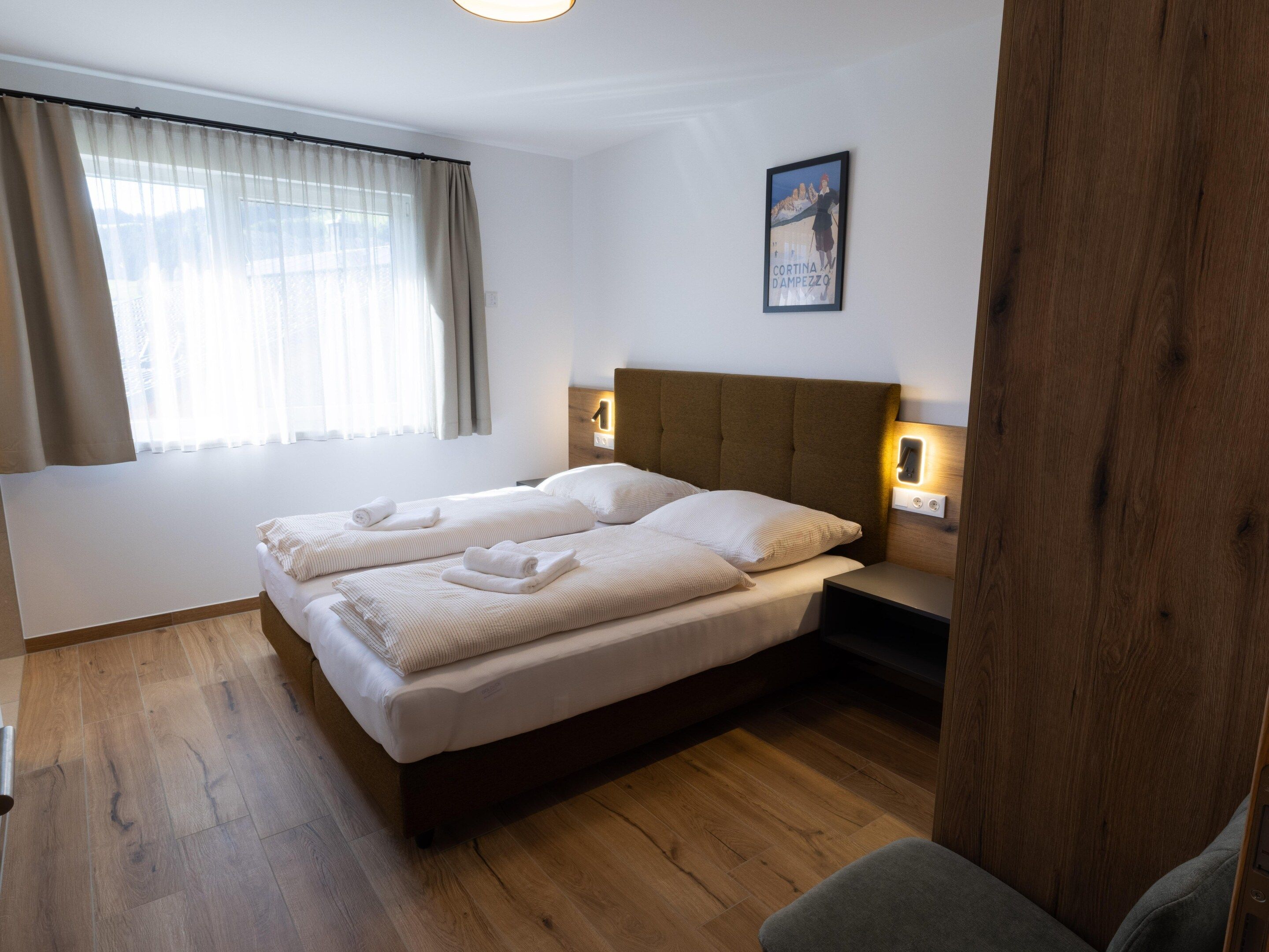 A double bed with two pillows and two rolled towels, a bedside table with a lamp, a framed picture on the wall, and a window with curtains.