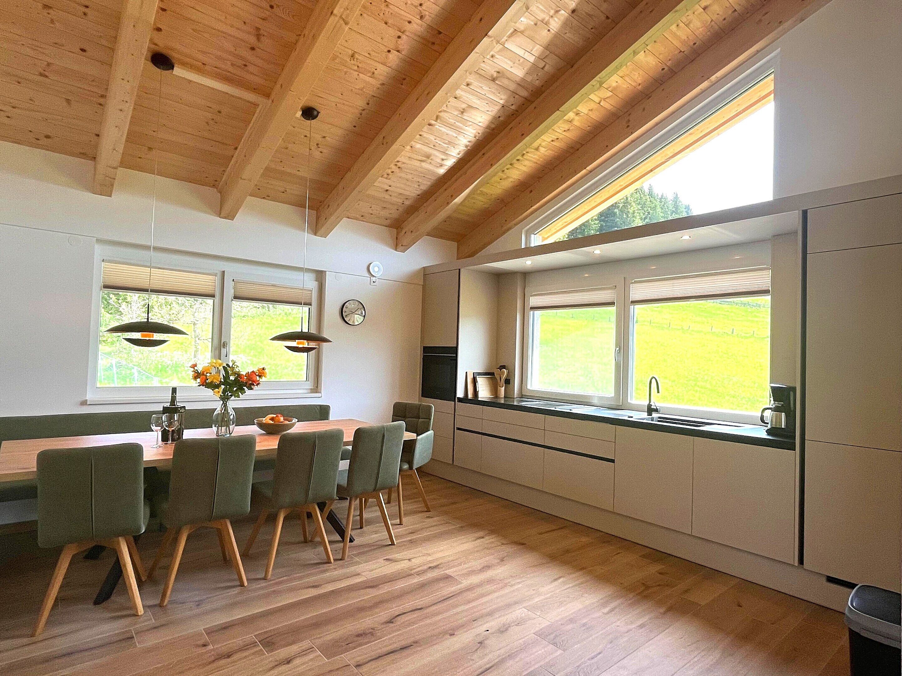 A modern kitchen with wooden beams, a dining area with chairs, a clock, and a view of the outdoors.