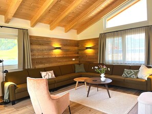 Un salon chaleureux avec un plafond en bois, un canapé marron, un fauteuil rose, une table basse ronde et un vase rempli de fleurs.
