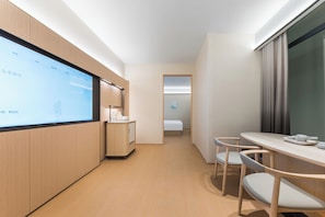 Moderni hotellihuone, jossa on suuri näyttö, jolta näkyy tietoa, puinen työpöytä tuoleineen, sänky ja pieni keittokomero.