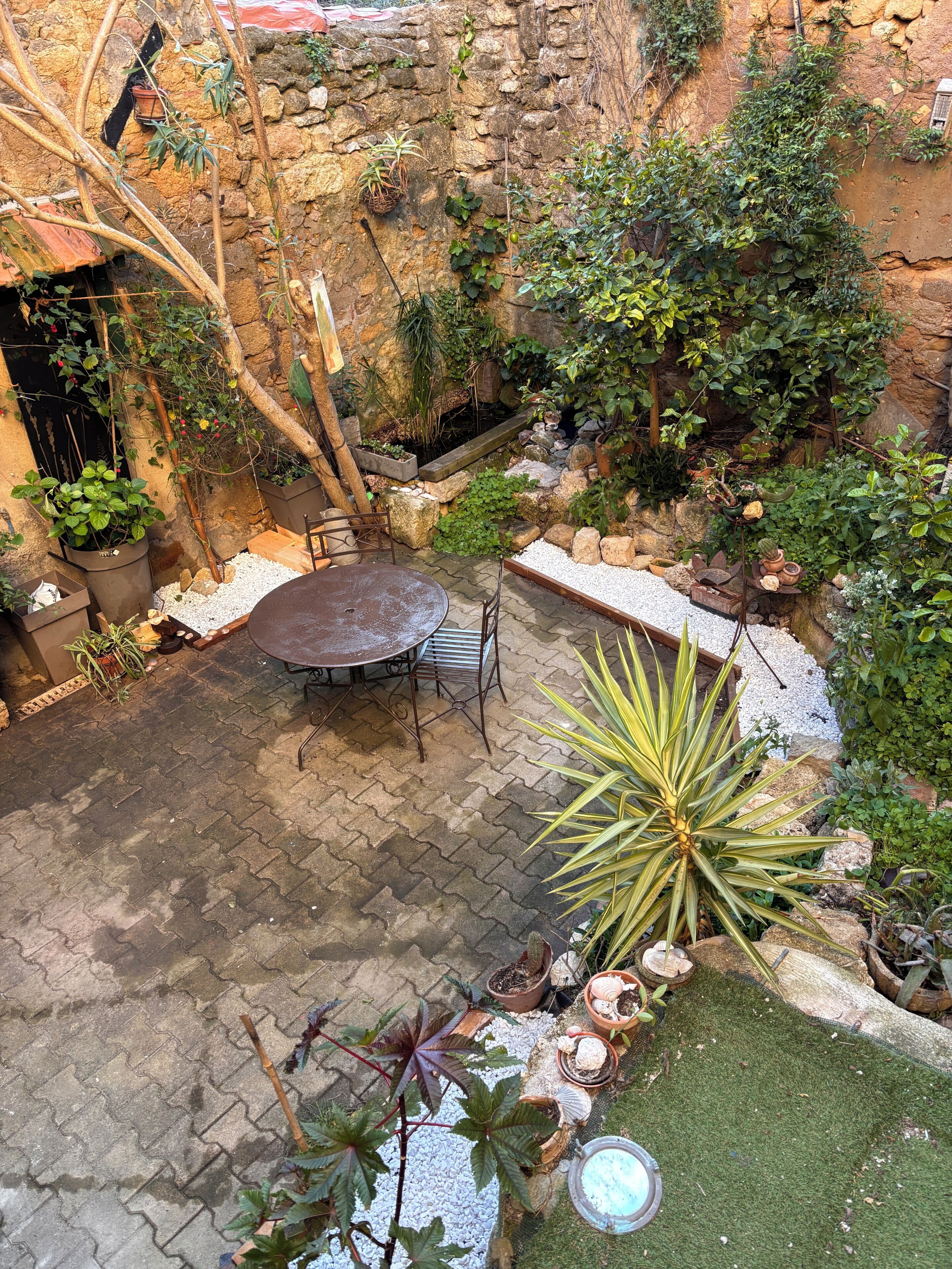 Un patio con pared de piedra, plantas en macetas, una mesa metálica y un pequeño jardín.