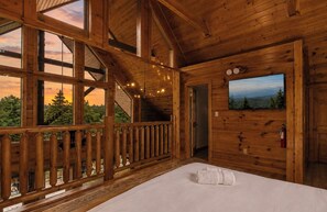 A wooden cabin room with a bed, a window with a view of trees, a door, and a wall-mounted picture. - Hot Tub, Pool Table & Arcade I Ridgetop View Cabin (Sevierville)