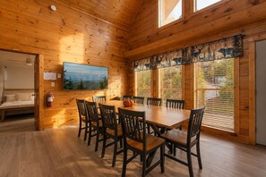 Interior de una cabaña de madera con una mesa de comedor, sillas, una cama y un gran ventanal con vistas a árboles.