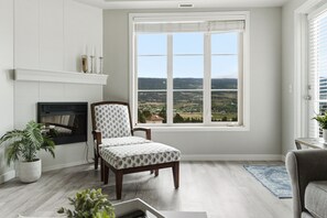 A living room with a fireplace, a large window with a view, a patterned armchair, and a potted plant. - Pinnacle Pointe | Valley-View Golf Escape (Kelowna)