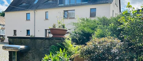 Una casa de dos pisos con techo plano, balcón y jardín con diversas plantas.