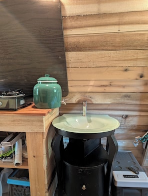 A compact kitchen with a stove, a green pot, a sink, and a trash bin. - Tranquil Glamping Cabin in Athens (Athens)
