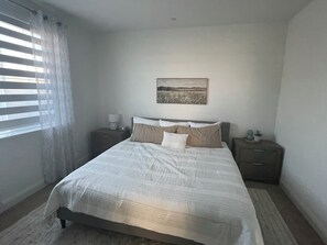A bedroom with a bed, two bedside tables, a nightstand, a window with blinds, and a wall-mounted picture.