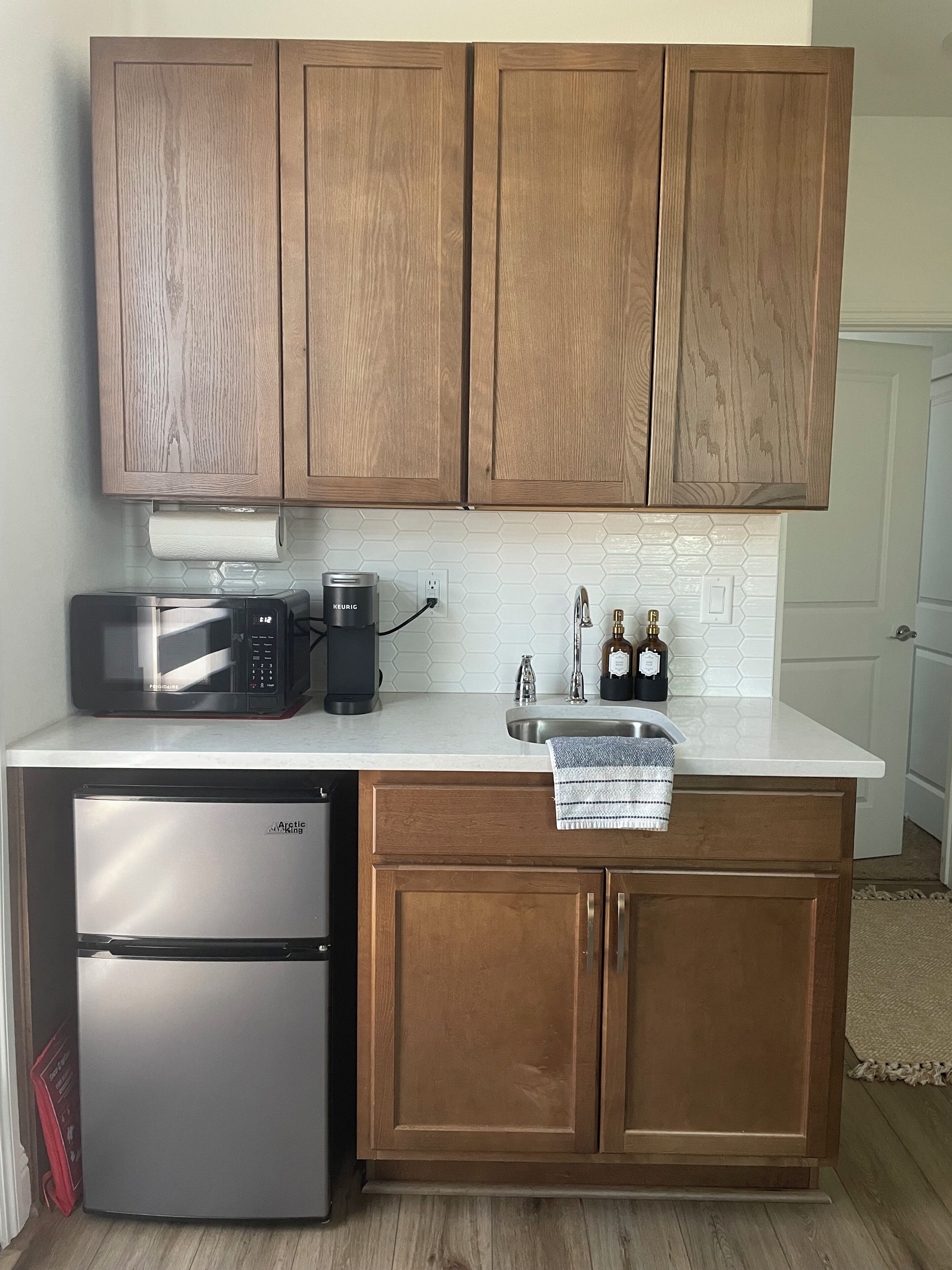 A compact kitchen with wooden cabinets, a stainless steel refrigerator, a microwave, and a coffee maker.