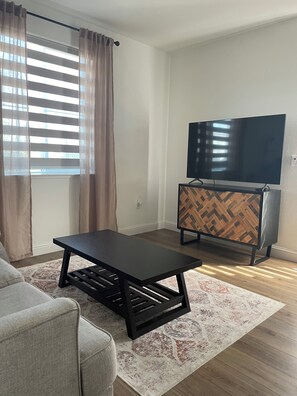 A living room with a sofa, a coffee table, a TV on a wooden cabinet, and a window with curtains. - Cute Boho Guest Suite | Private Entry | Great Location (Roseville)