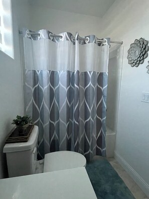 A bathroom with a shower curtain, a toilet, a small table with a plant, and a decorative wall piece. - Cute Boho Guest Suite | Private Entry | Great Location (Roseville)