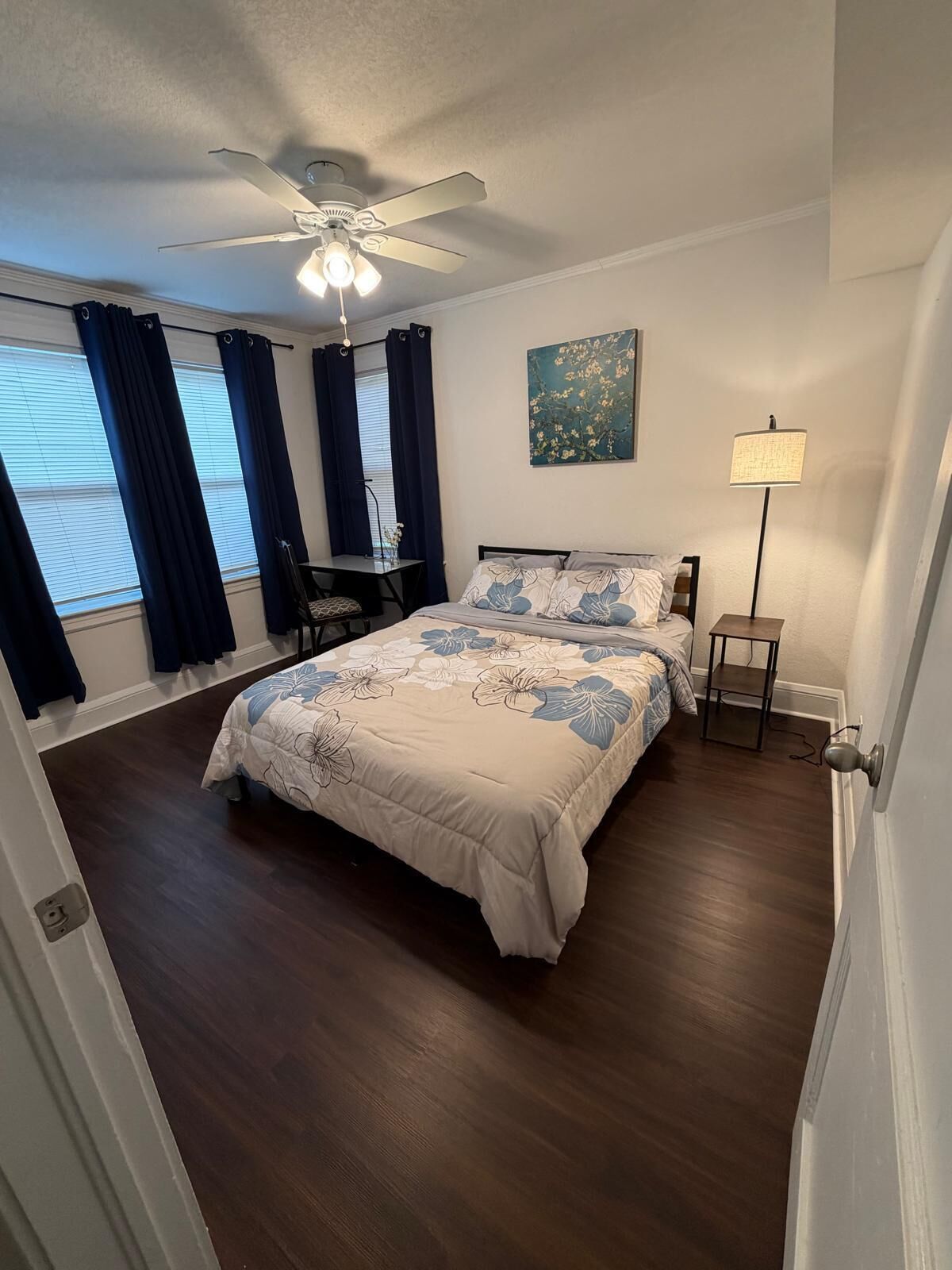 A bedroom with a bed, a desk, a chair, a standing lamp, and a ceiling fan.