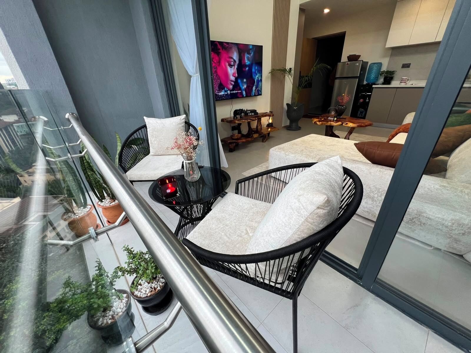 A balcony with a glass railing, two black wicker chairs with white cushions, a small table with a candle, and potted plants.