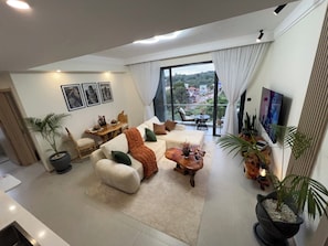 Ein großzügiger Wohnbereich mit einer Sofagarnitur, einem Couchtisch und einem Esstisch mit Stühlen. Es befinden sich Blumentöpfe, ein an der Wand montierter Fernseher und ein Balkon mit Blick auf die Stadt darin.