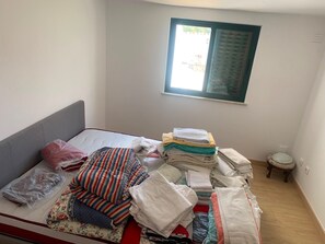 A small, single-bed room with a window, a small stool, and a pile of folded towels and blankets on the bed. - Sleeps4-familyfriendly-balcony (Lagoa)