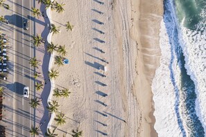 Eine Vogelperspektive zeigt einen Strand mit Palmreihen, eine Straße parallel zum Strand und Brandwellen, die auf die Küste branden.