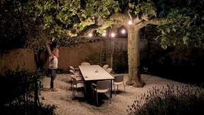 Zona d'exterior per dinar amb taula i caires, envoltada d'arbres i una paret de pedra.