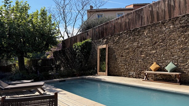Piscina amb terrassa de fusta, mur de pedra i una banca amb coixins.