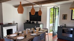 Zona de dinar amb taula de fusta preparada per a sis, envoltada per sis cadirees. Hi ha una llar de foc, cuina amb fogó i una finestra gran que ofereix vistes a vegetació exterior.
