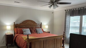Un dormitorio con una cama de madera, dos mesitas de noche con lámparas, un ventilador de techo y una ventana con cortinas.