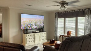 Una sala de estar con televisor de pantalla plana, ventilador de techo, sofá, mesa de centro y ventana con persianas.