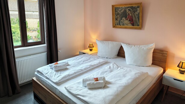 Een hotelkamer met een groot bed, twee nachtkastjes, een raam met gordijnen, een radiator en een schilderij aan de muur.