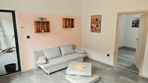 Een woonkamer met een grijze bank, een salontafel, houten planken en een deur naar een andere kamer.