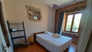 A bedroom with a bed, a shelf with a lamp, a window with curtains, and a wall-mounted picture frame. - Gite 2 Moulin SAINT ETIENNE (Bédarieux)