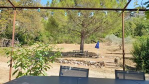 A covered outdoor seating area with a table and chairs, surrounded by trees and a stone wall. - Gite 2 Moulin SAINT ETIENNE (Bédarieux)