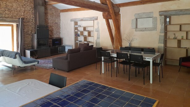 A rustic interior with exposed wooden beams, a stone wall, a fireplace, a sofa, a dining table with chairs, and a tiled floor. - Gite 2 Moulin SAINT ETIENNE (Bédarieux)