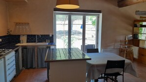 A kitchen with a central island, a dining table with chairs, a hanging light fixture, and a view through a glass door. - Gite 2 Moulin SAINT ETIENNE (Bédarieux)