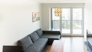 Un soggiorno moderno con un divano ad angolo grigio, un tavolino in legno, un portatile su una scrivania e un'ampia finestra con vista sugli edifici.