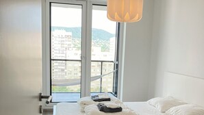 Una camera da letto con un letto, biancheria bianca, un lampadario a sospensione, una finestra vista edifici e un pavimento in legno.