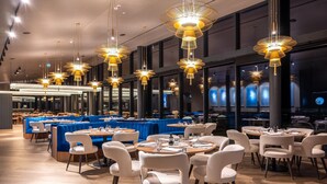 En restaurant med flere spiseborde, stole og hængende pendellamper.