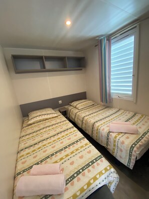 A room with two single beds, a window with blinds, a shelf above the beds, and a ceiling light. - Mobile home near beach Le Pradet (Var, 83) (Le Pradet)