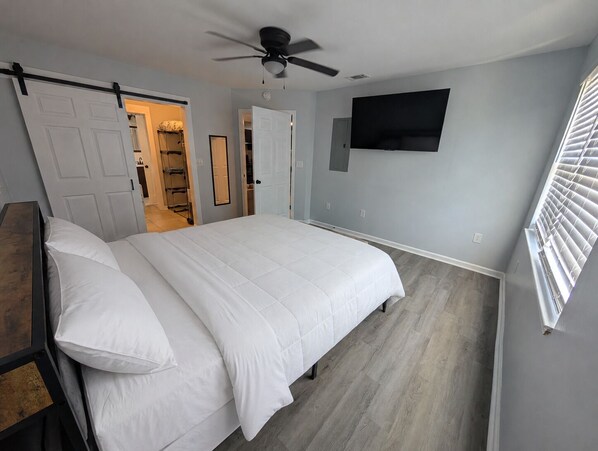 Une chambre à coucher avec un lit, un ventilateur de plafond, un téléviseur fixé au mur et une fenêtre avec des stores.