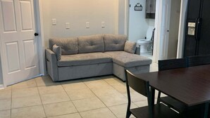 A room with a sectional sofa, a dining table with chairs, a ceiling fan, and a kitchen area visible through an open doorway. - The Navarre Nook at The Spires - Near Navarre Beach & Pier (Navarre)