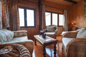 Une pièce aux murs lambrés, avec un canapé en osier, un fauteuil, une petite table sur laquelle se trouve une lampe, et de grandes fenêtres.
